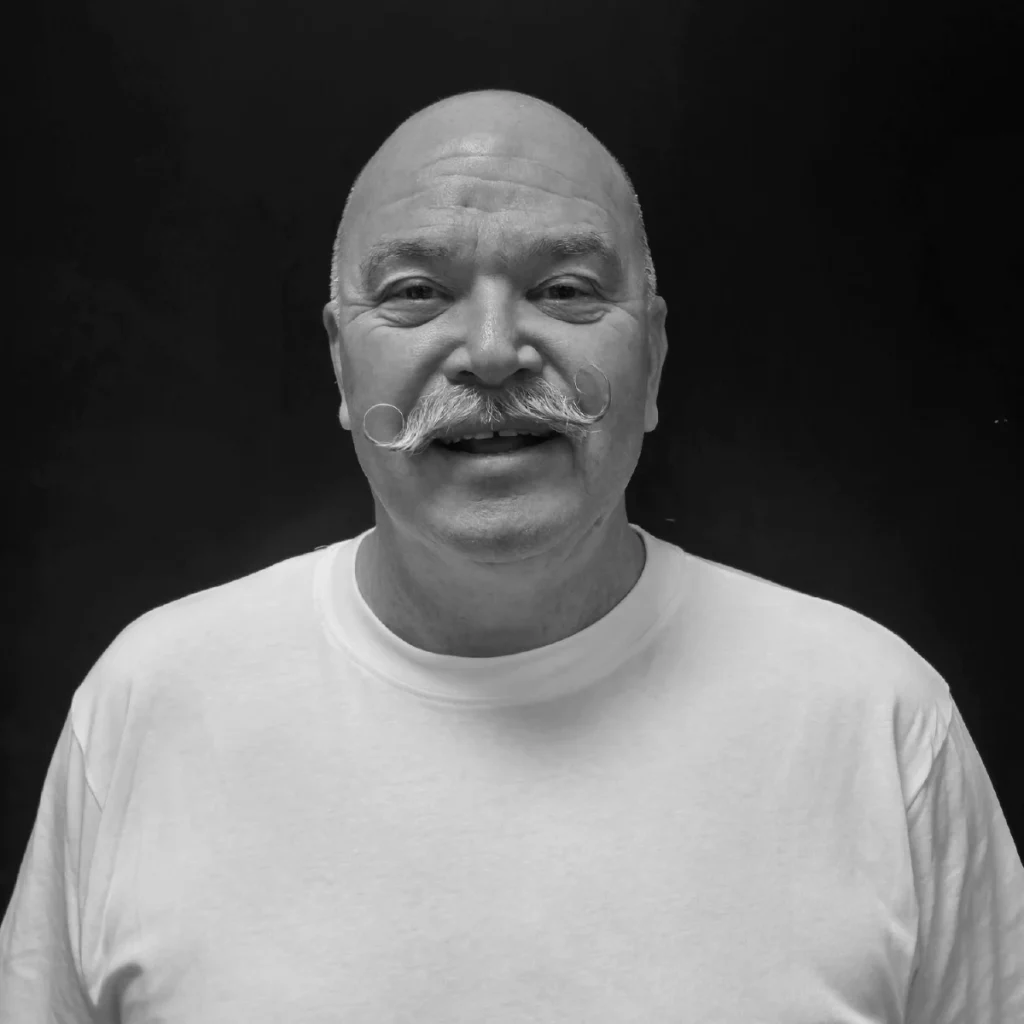 En voksen mann uten hår og med krøllet bart, iført enkel hvit T-skjorte, smiler foran en nøytral, mørk bakgrunn.