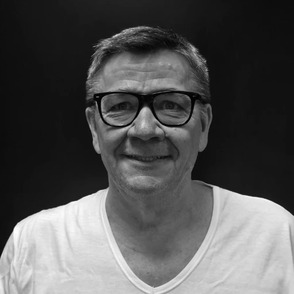 Erfaren mann med kort hår og briller i hvit skjorte, smilende foran nøytral bakgrunn, fremstår profesjonell og tillitsvekkende.
